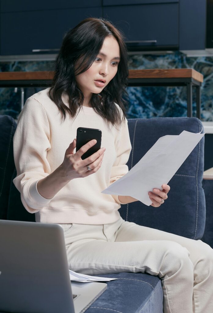 pexels-photo-6963013-6963013 A young woman uses her smartphone and laptop to manage finances, appearing focused and concerned.